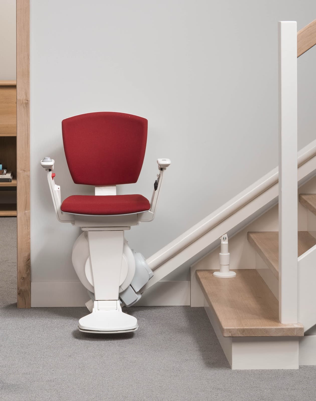 A modern stairlift with a red seat parked at the bottom of a light wooden staircase