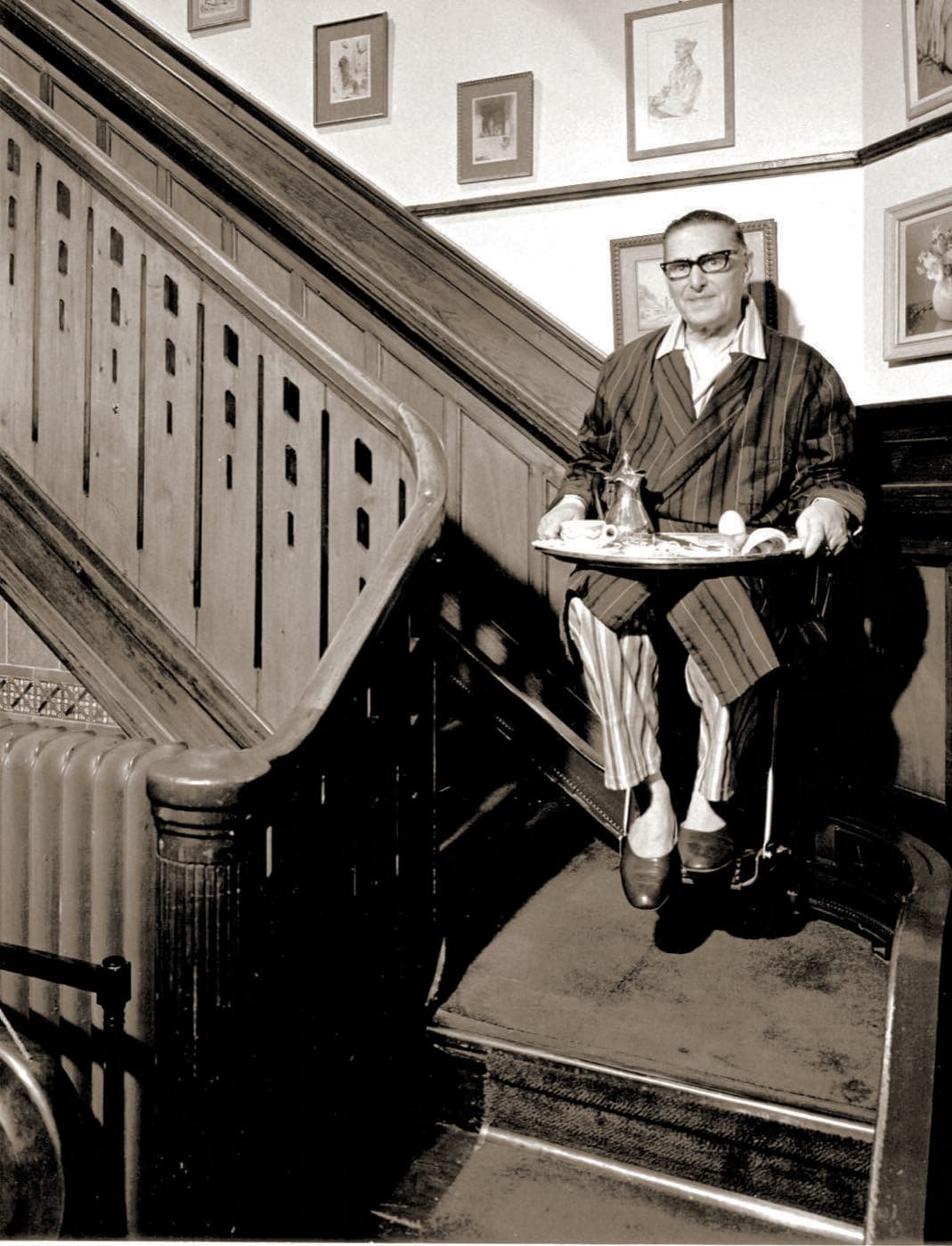 A vintage mid-century photograph of a man riding an early stairlift while holding a breakfast tray