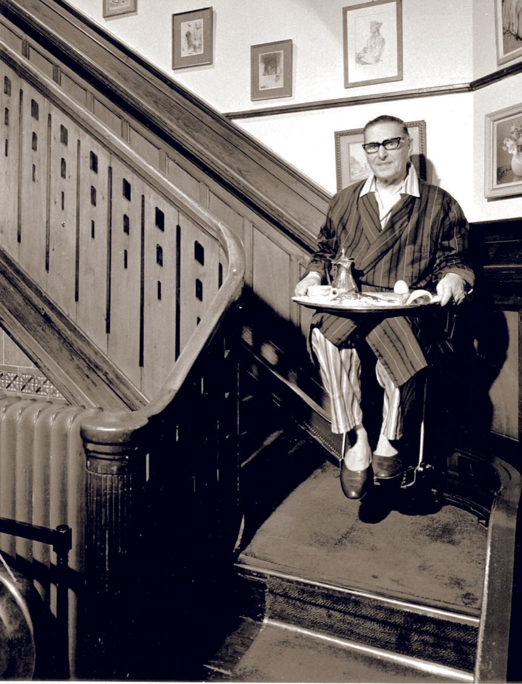 A vintage mid-century photograph of a man riding an early stairlift while holding a breakfast tray