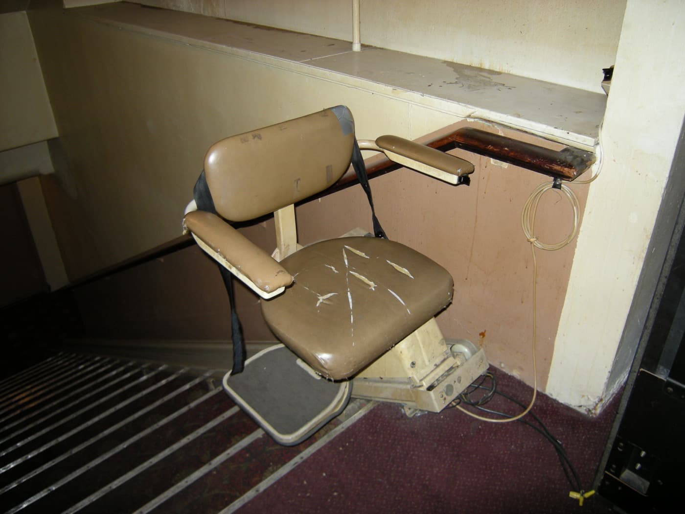 An old, heavily worn stairlift with cracked upholstery at the bottom of a staircase